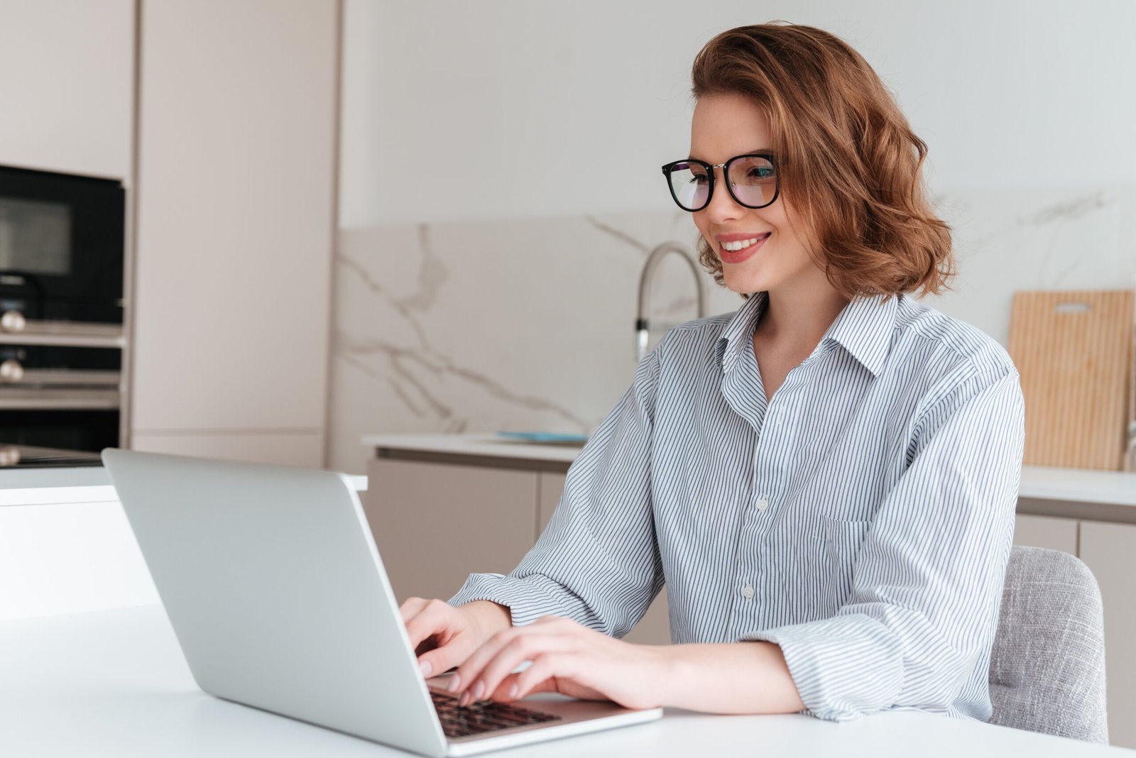 Elegant smiling woman in glasses and striped shirt using laptop Elegant smiling woman in glasses and striped shirt using laptop computer while siting at table in kitchen